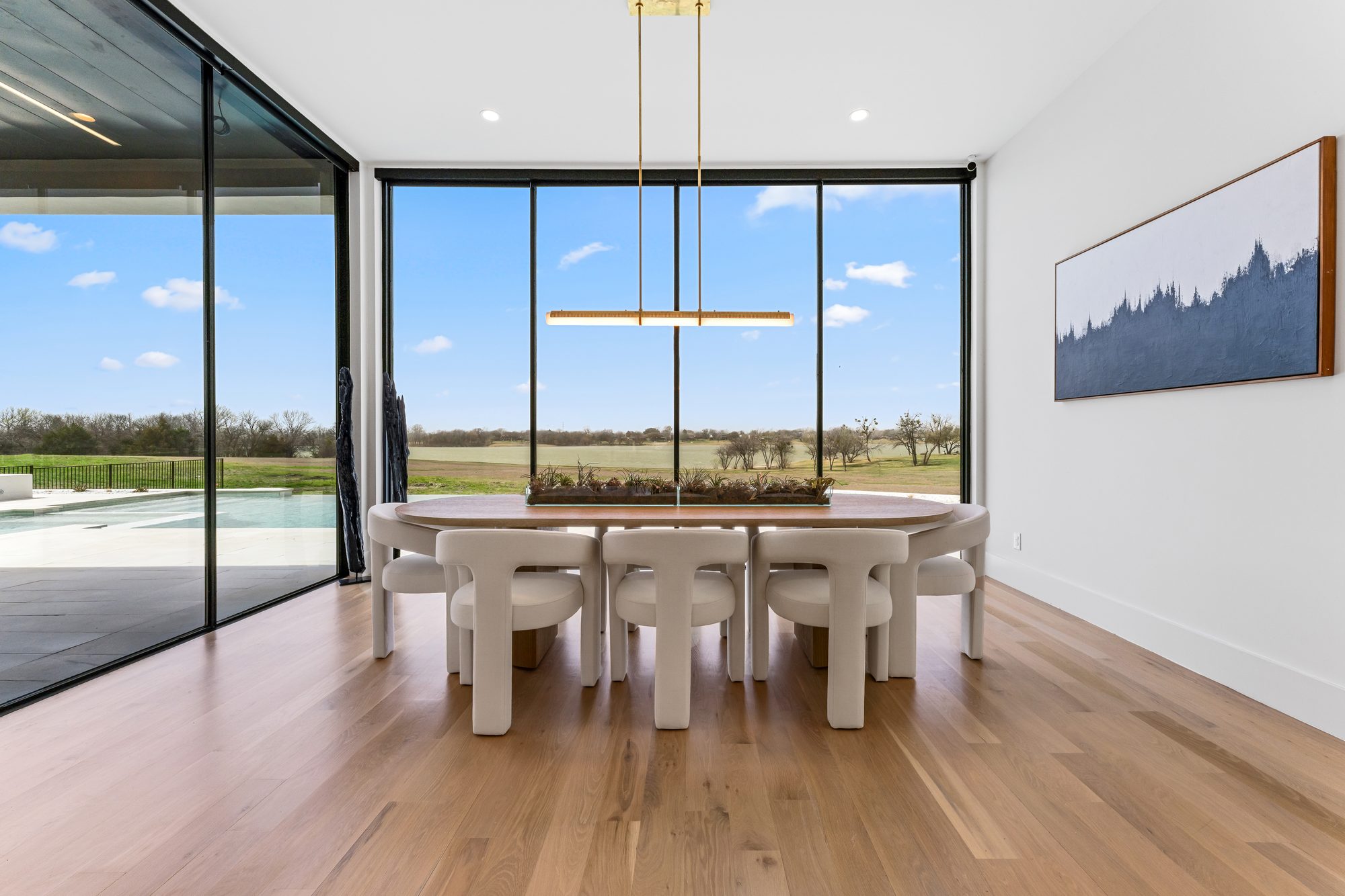 Dining area with a linear chandelier and walls of glass toward the pool and lawn